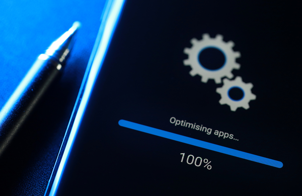 An Android phone displaying the update screen saying 'Optimising Apps' with two cogs spinning. The device is on a blue background with a pen next to it. 
