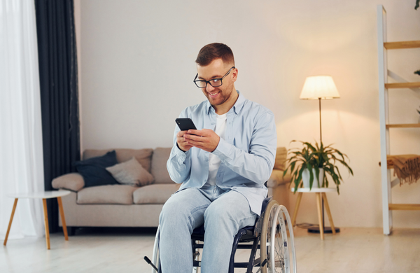 A man in a wheelchair wearing a light blue shirt, jeans and black thick rimmed glasses in a lounge room setting. The man is holding a phone and is looking down smiling at it. 