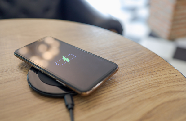 A smartphone wirelessly charging on a wooden table. The smartphone screen is displaying the charging symbol of a battery and lightening bolt through it. 