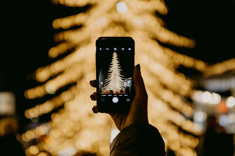 A hand holding up a phone taking a picture of a Christmas tree glowing in golden lights. 