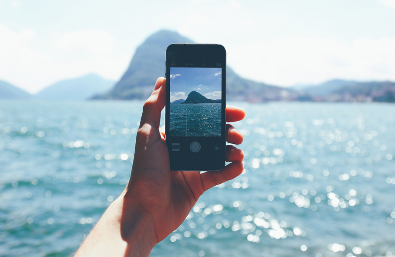 A hand holding a smartphone with the camera open, viewing over the ocean and islands