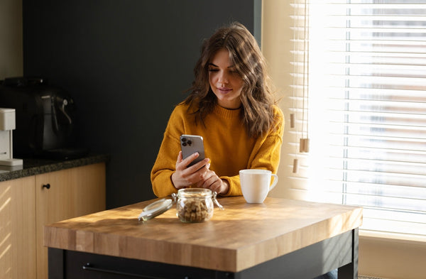 A woman sitting at a kitchen bench with the morning sun shining in while she looks down at her phone.