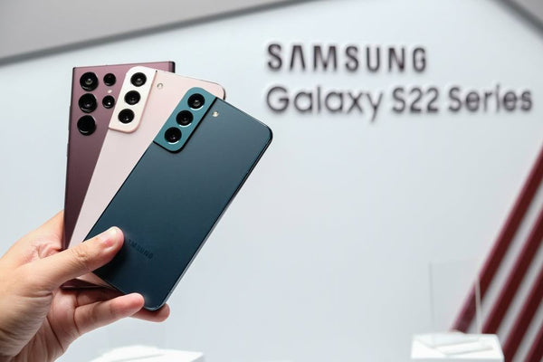 Caucasian hand holding three Samsung Galaxy S22 phones: one burgundy, one pink and one dark blue