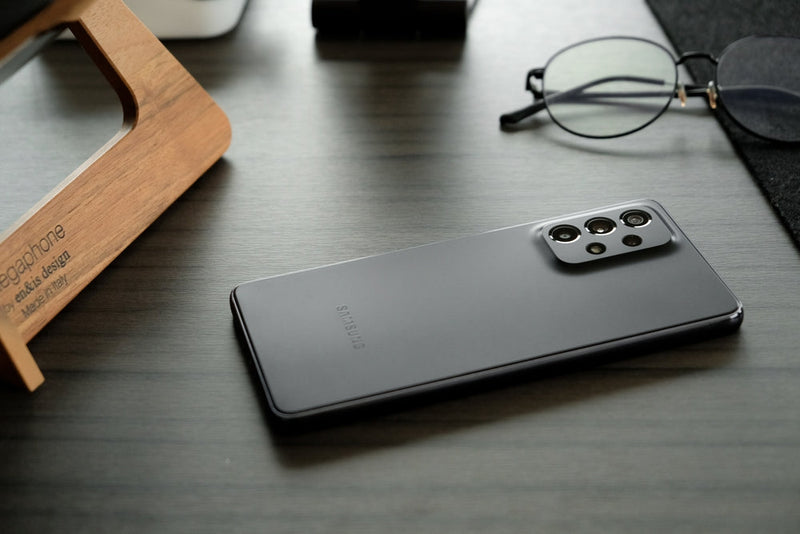 A black Samsung Galaxy A series phone screen down on a desk next to reading glasses.