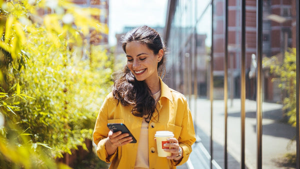 A woman walking along in public holding a coffee a looking down at her phone smiling. 