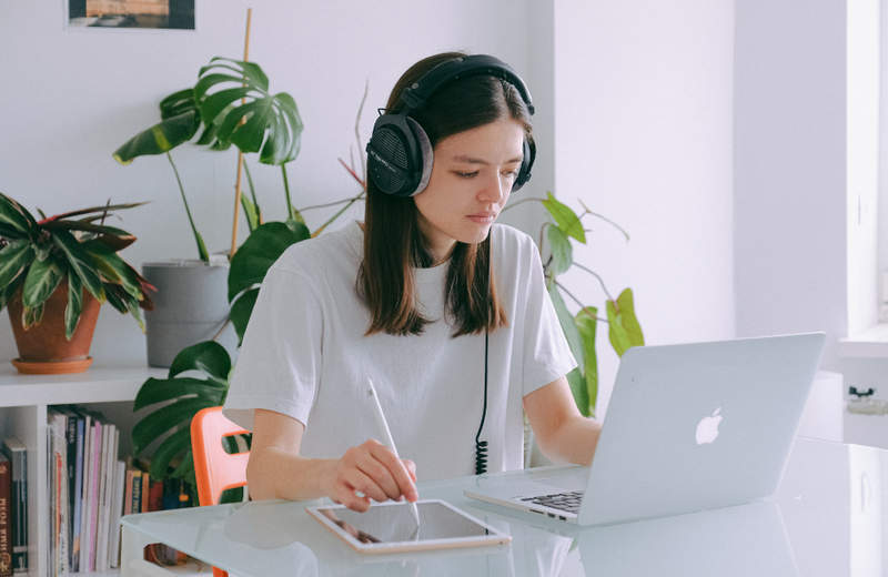 A student sitting in a home office with a MacBook and iPad on the table. The student is wearing headphones and there are plants and books in the background. 