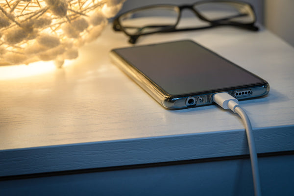 A phone charging on a bedside table next to a pair of glasses. 