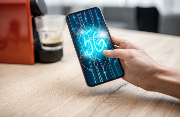 A hand holding a phone in front of a pod coffee machine. The phone has 5G technology graphics on the screen. 