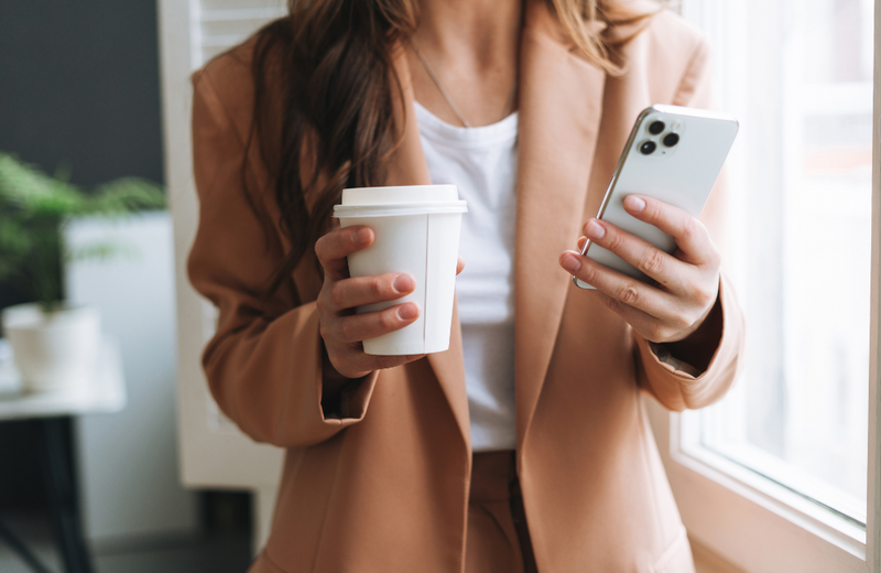 A corporate woman in an office space holding a takeaway coffee cup in one hand and a white iPhone in the other. 