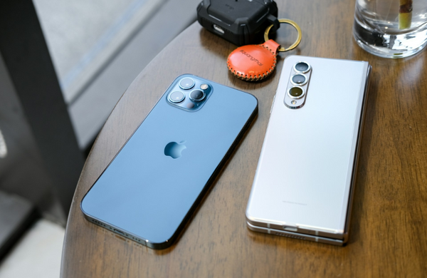 An icy blue iPhone next to a white silver Samsung Galaxy Fold phone facedown on a wooden table.