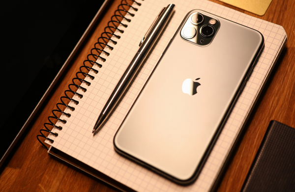 A gold iPhone 15 next to a gold pen on top of a notepad on a wooden desk. 