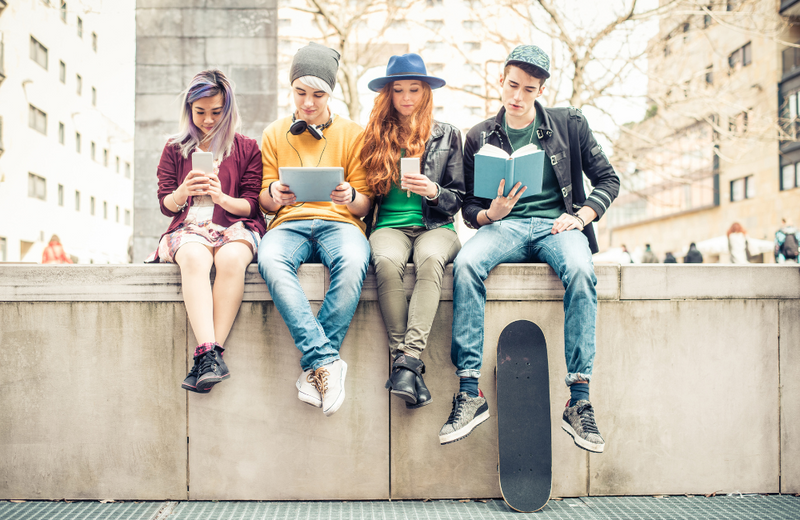 Four teenagers sitting on a wall all looking down at different items: a phone, a table, an iPhone, and a book.