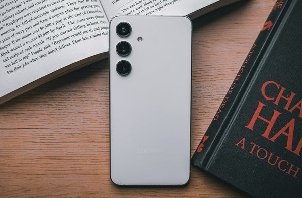 A white Samsung Galaxy S24 face down with the top end propped up by an open book. Next to it, there is a black closed book with red letting on the cover. The books and phones are resting on a wooden surface.  