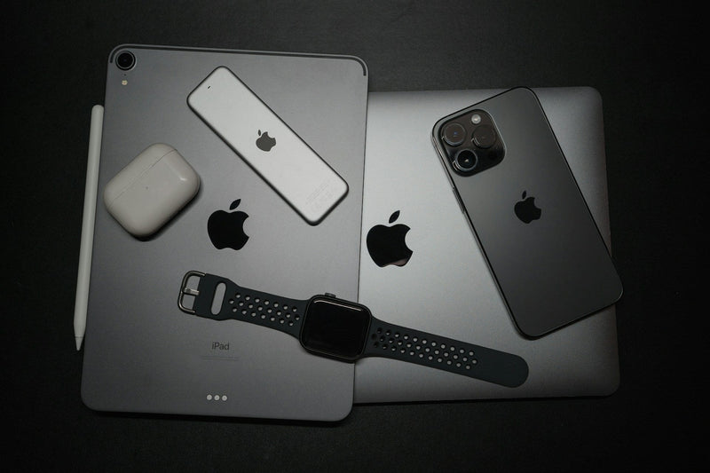 A MacBook, iPad, iPhone, Air Pods, Apple Watch and other Apple aaccories stacked up on a black background. All the devices are a range of grey or white.