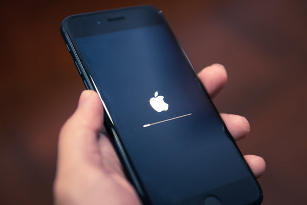 A hand holding a black iPhone with the Apple symbol displayed on the screen with a loading bar beneath it. 