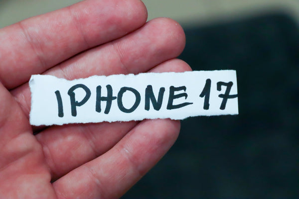 A hand holding a piece of paper saying iPhone 17 in marker pen.