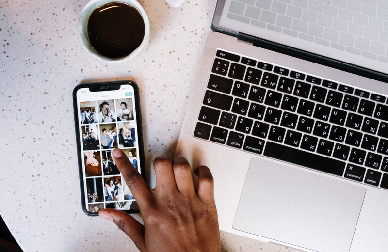 A phone displaying the image gallery on its screen next to a laptop and coffee. There is a hand with a finger poised over the screen.