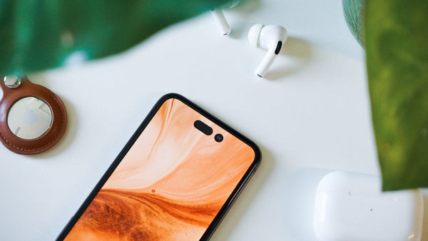 A black iPhone 14 with an orange display next to Air Pods with a plant unfocused in the forefront